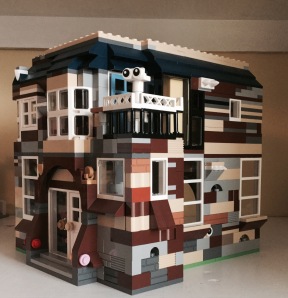 A two-storey house built of brown, grey and beige Lego bricks. A simple 'ghost' made of white bricks stands in an upstairs balcony.