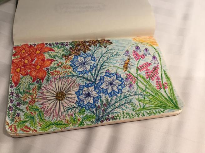 A paper Moleskine sketchpad, opened out to show a colourful pen-drawn scene, landscape orientation, featuring flowers of various shapes, colours and sizes, foliage, and a honeybee. The sun shines in the background, in the top-right corner of the drawing.